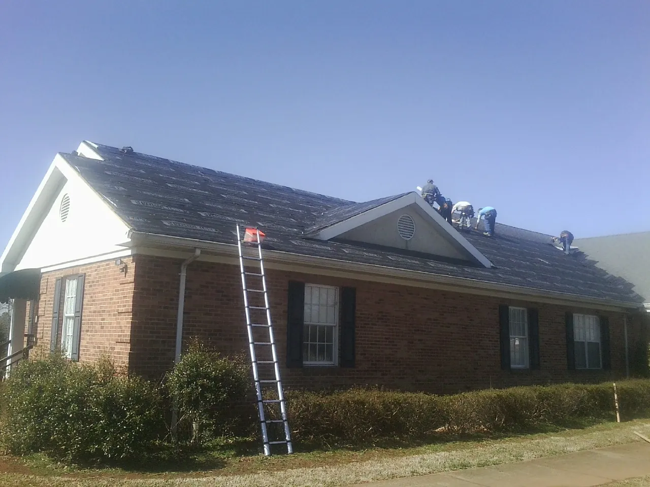 Crew tearing shingles off a brick ranch roof with a ladder set — Metal Roof Repair work by our crew in Burns